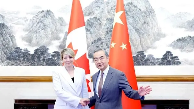 Mélanie Joly shakes hands with Wang Yi in front of Canadian and Chinese flags.