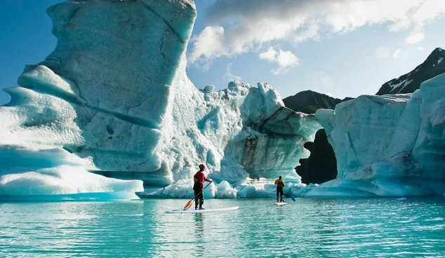 Two adults on stand up paddle board (SUP) observe hole melted in iceberg on Bear Lake in Kenai Fjords National Park, Alaska.